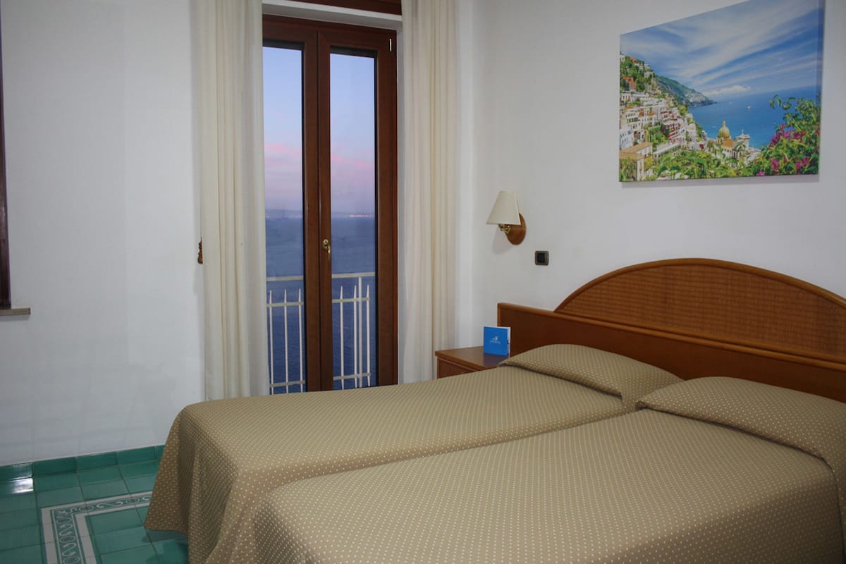 Ein Hotelzimmer mit Bett, Nachttisch, einem Gemälde und Meerblick durch eine Tür.