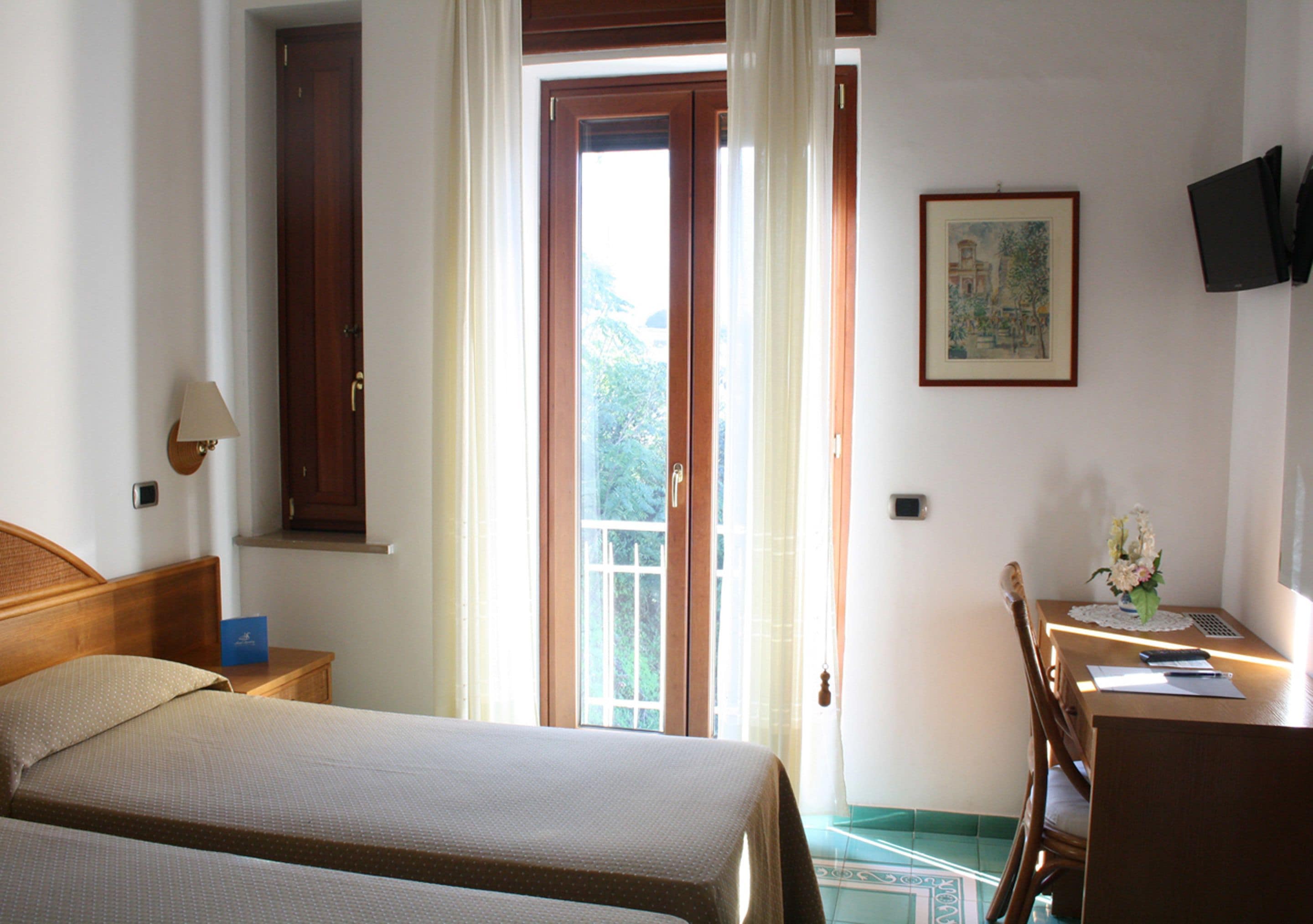 A hotel room with a bed, a desk with a chair, a small table with a vase, a painting on the wall, and a door leading to a balcony.