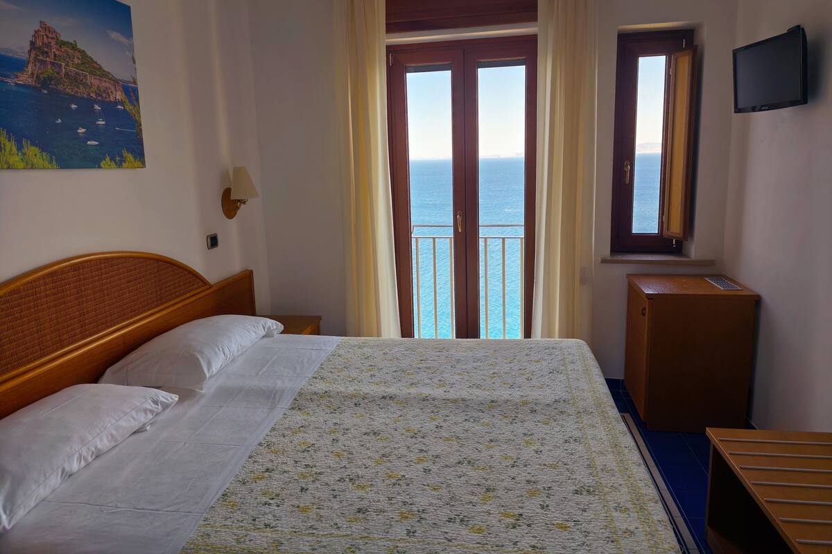 Ein Hotelzimmer mit Bett, Nachttisch, Lampe, Fernseher, Fenster mit Meerblick und einem Bild an der Wand.