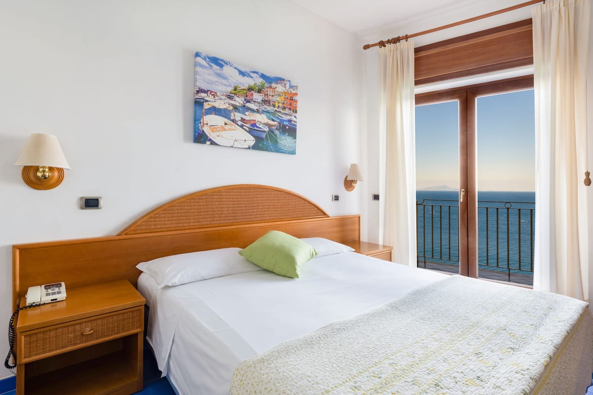 Ein Hotelzimmer mit einem hölzernen Bett, einem Gemälde einer Küstenlandschaft, einem Nachttischchen mit Telefon und Blick auf das Meer vom Balkon.