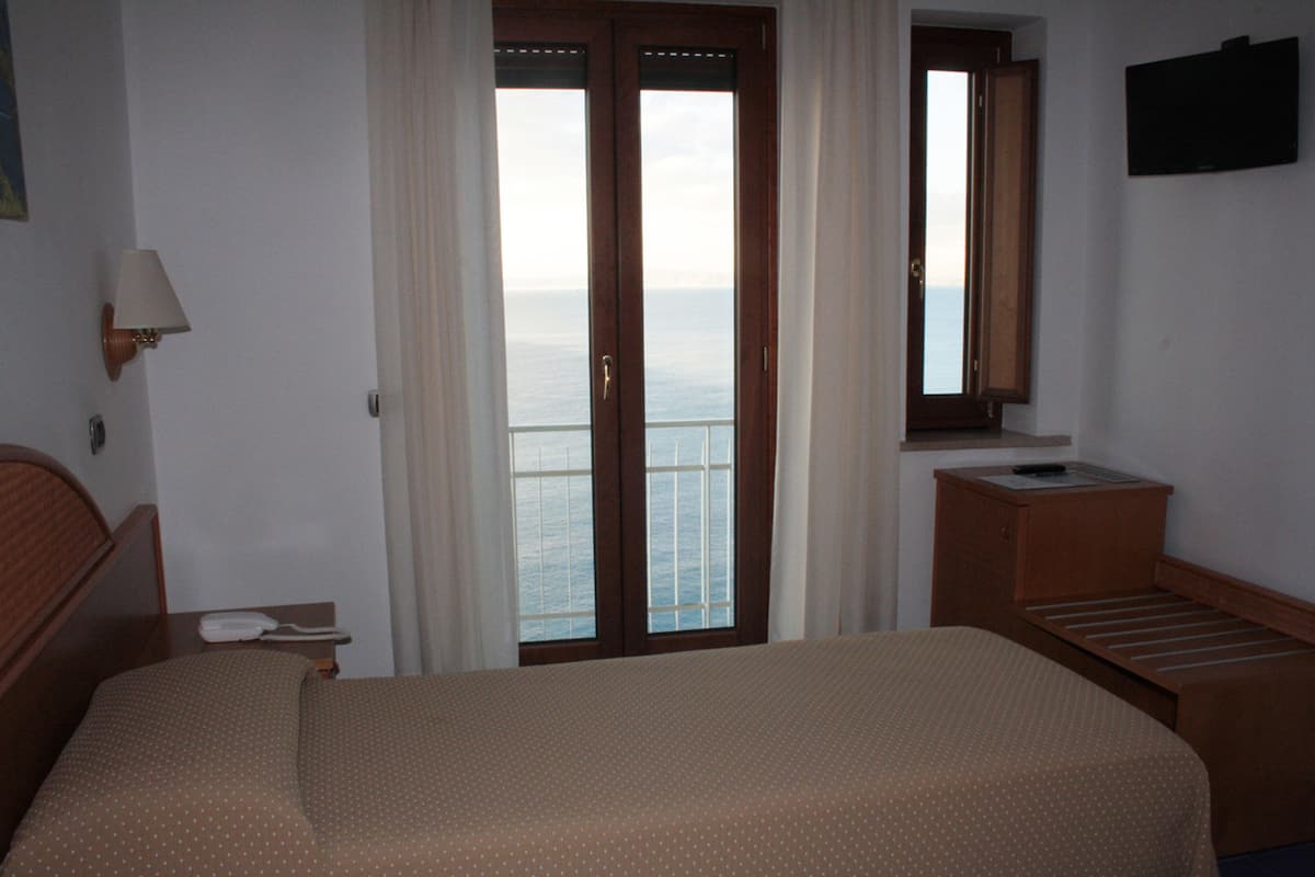 Ein Hotelzimmer mit Bett, Nachttisch, Lampe, Fenster mit Meerblick und kleinem Holztisch.