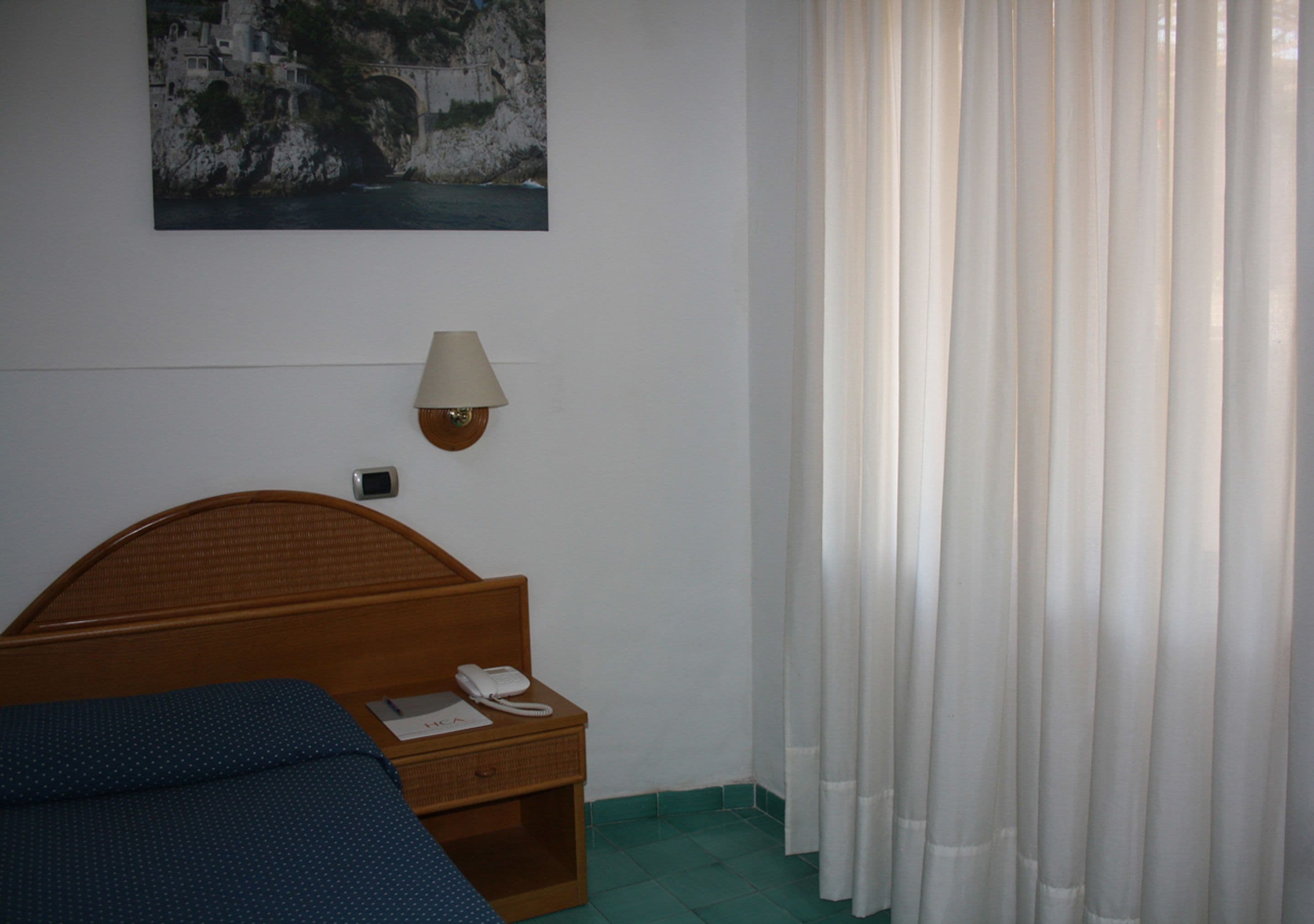 Habitación de hotel con una cama, una mesita de noche con lámpara y teléfono, un cuadro en la pared y una ventana con cortinas.