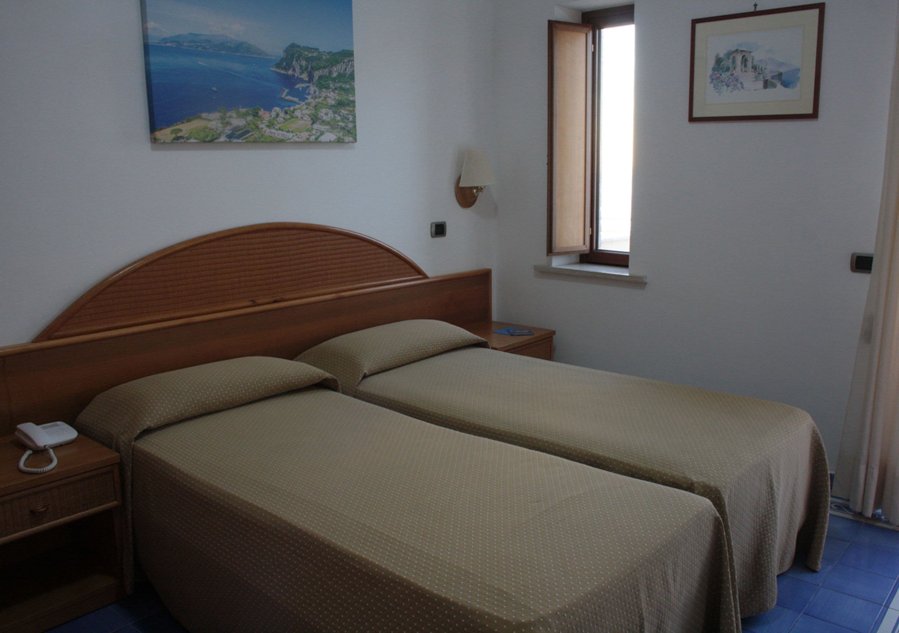 Ein Zimmer mit zwei Betten, einem Nachttisch mit Telefon, einem Fenster und Wandbildern.