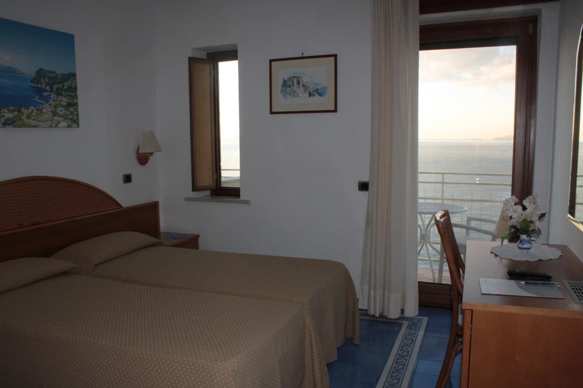 Ein Hotelzimmer mit einem Bett, einem Schreibtisch mit Blumen, einem Sessel, einem Fenster mit Meerblick und zwei gerahmten Bildern an der Wand.