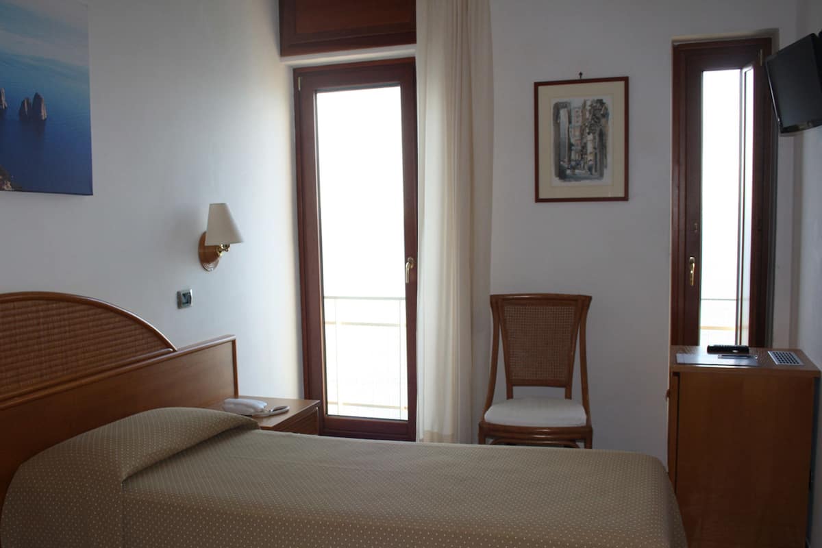 Ein Hotelzimmer mit Bett, Sessel, Schreibtisch mit Computer, Lampe und einem Bild an der Wand.