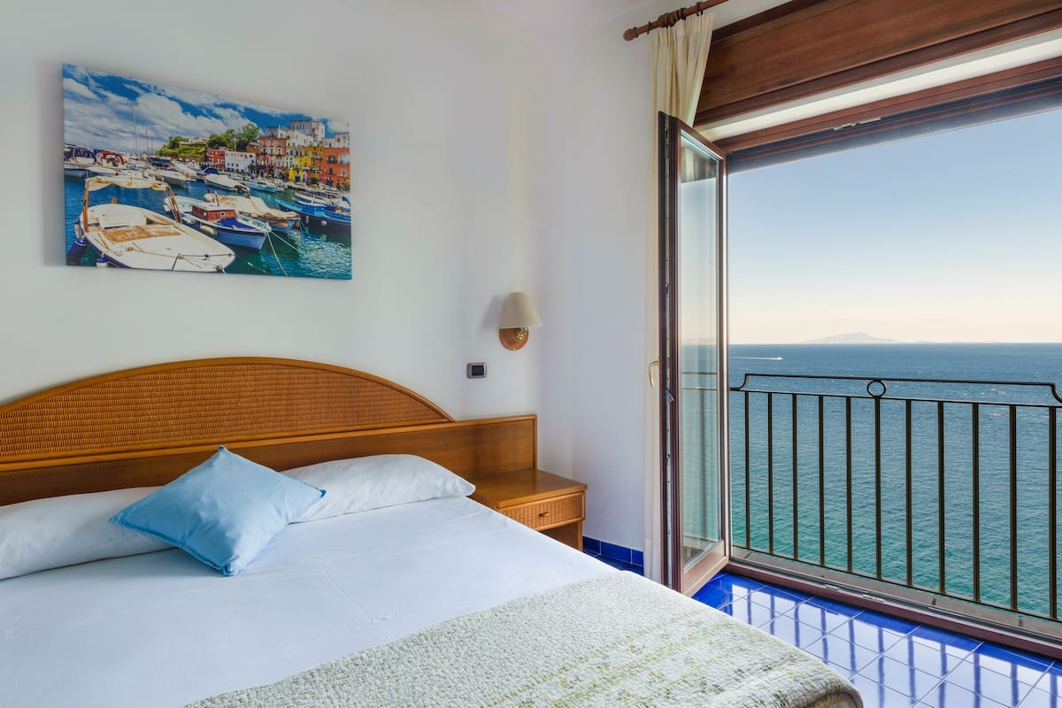 Ein Hotelzimmer mit einem Bett, einem Nachttisch, einer Lampe, einem Gemälde eines Hafens und einem Balkon mit Meerblick.