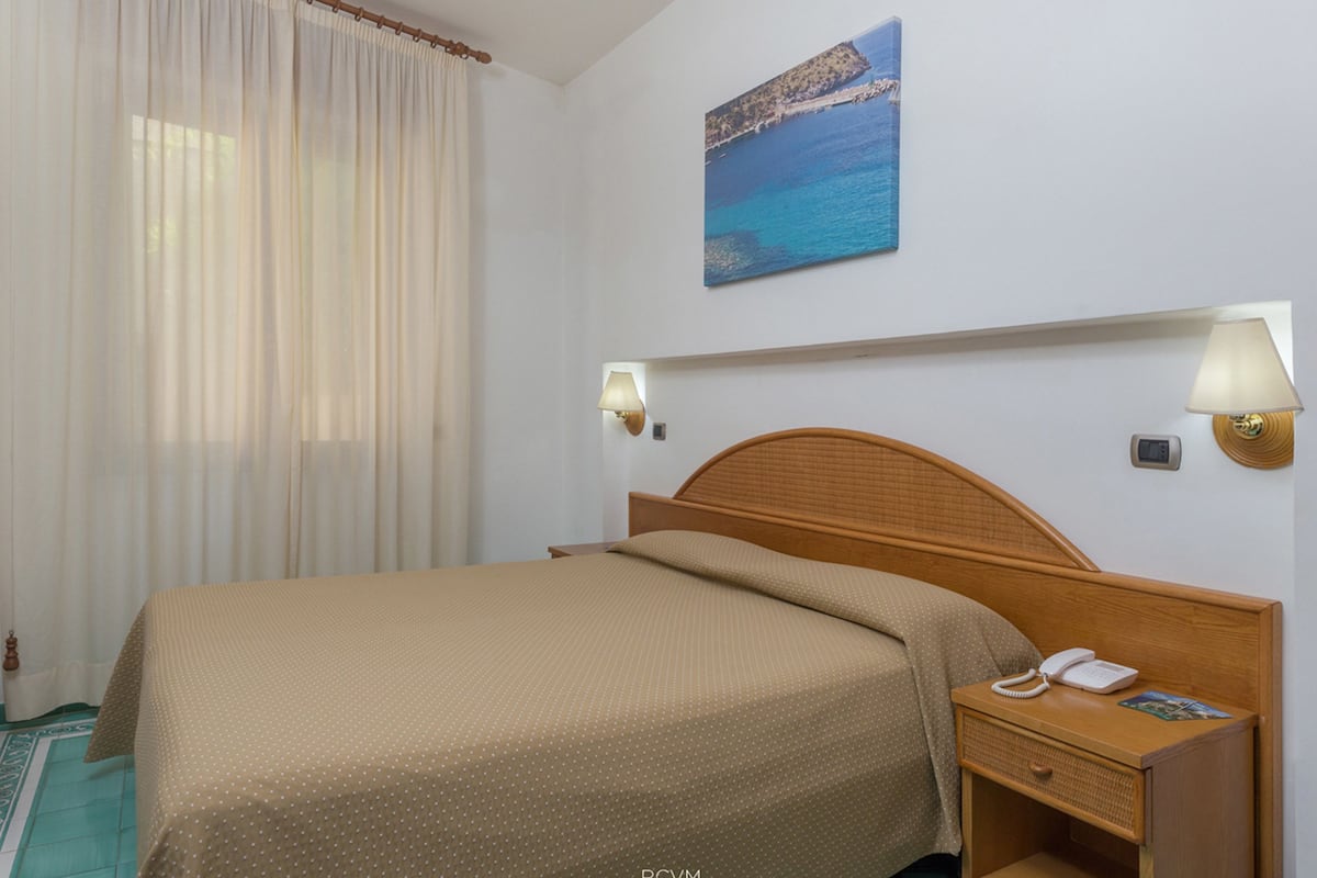 Ein Hotelzimmer mit Bett, Nachttisch mit Lampe, einem Bild mit Strandmotiv und einem Fenster mit Vorhängen.