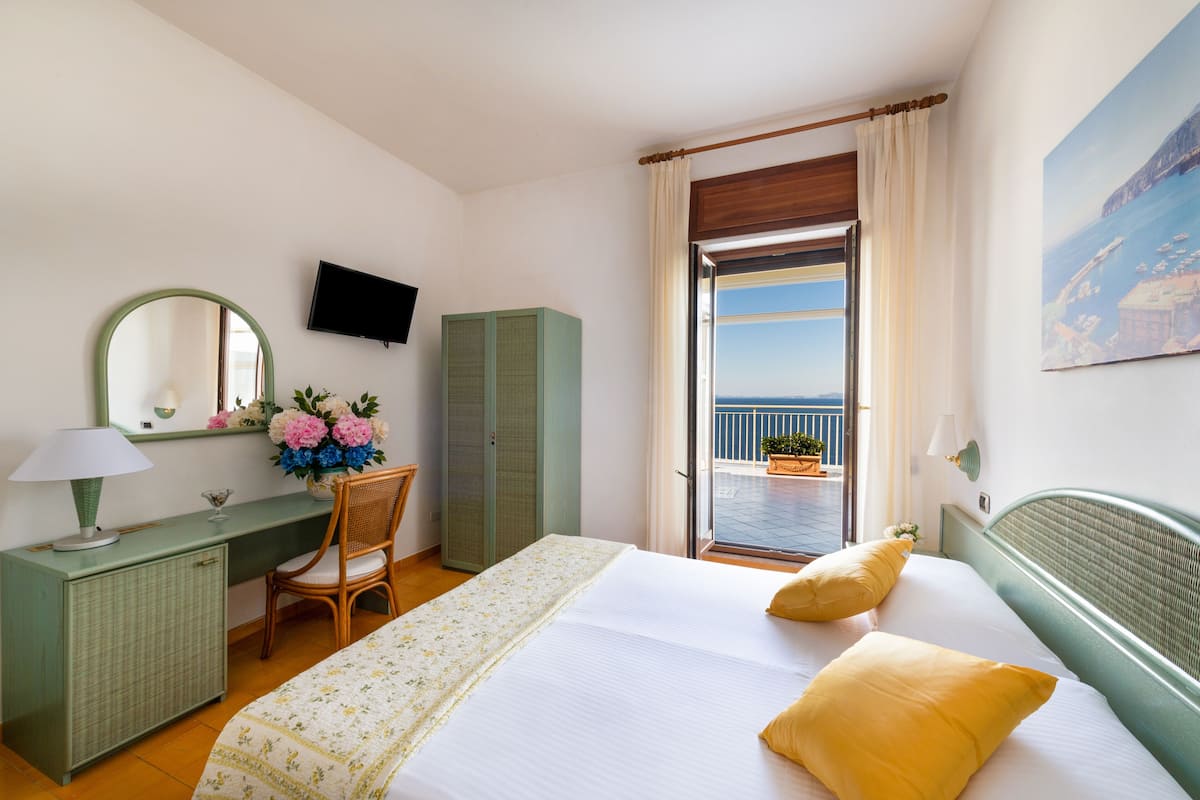 Ein Hotelzimmer mit Bett, Schreibtisch, Stuhl, Fernseher, einem Fenster mit Meerblick und einem Balkon mit Topfpflanzen.