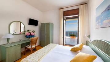 Une chambre d’hôtel comprenant un lit, un bureau, une chaise, une télévision, une fenêtre donnant sur la mer et un balcon agrémenté de plantes en pot.