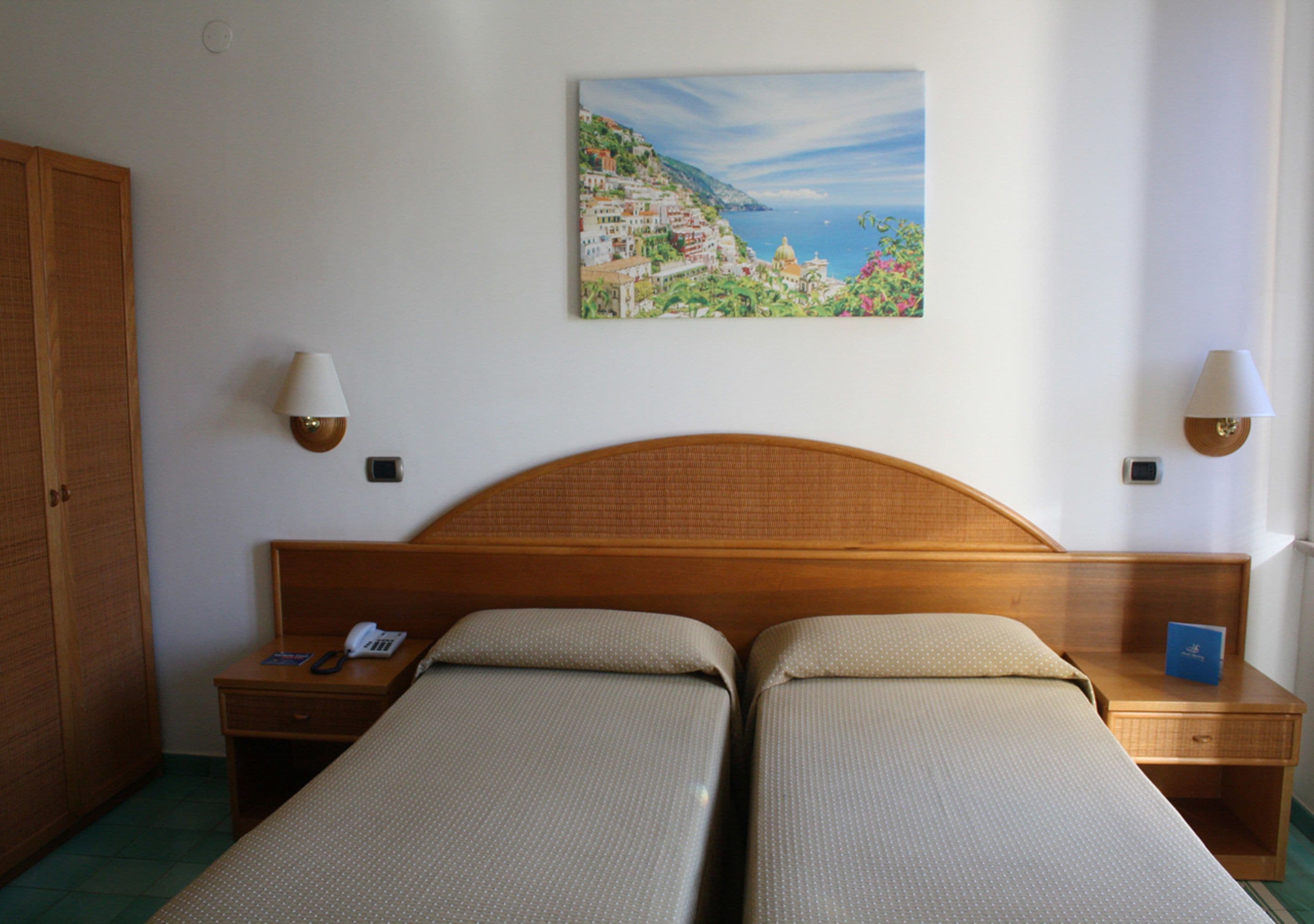 A hotel room with a wooden headboard, two single beds, a painting of a coastal scene, and two bedside lamps.