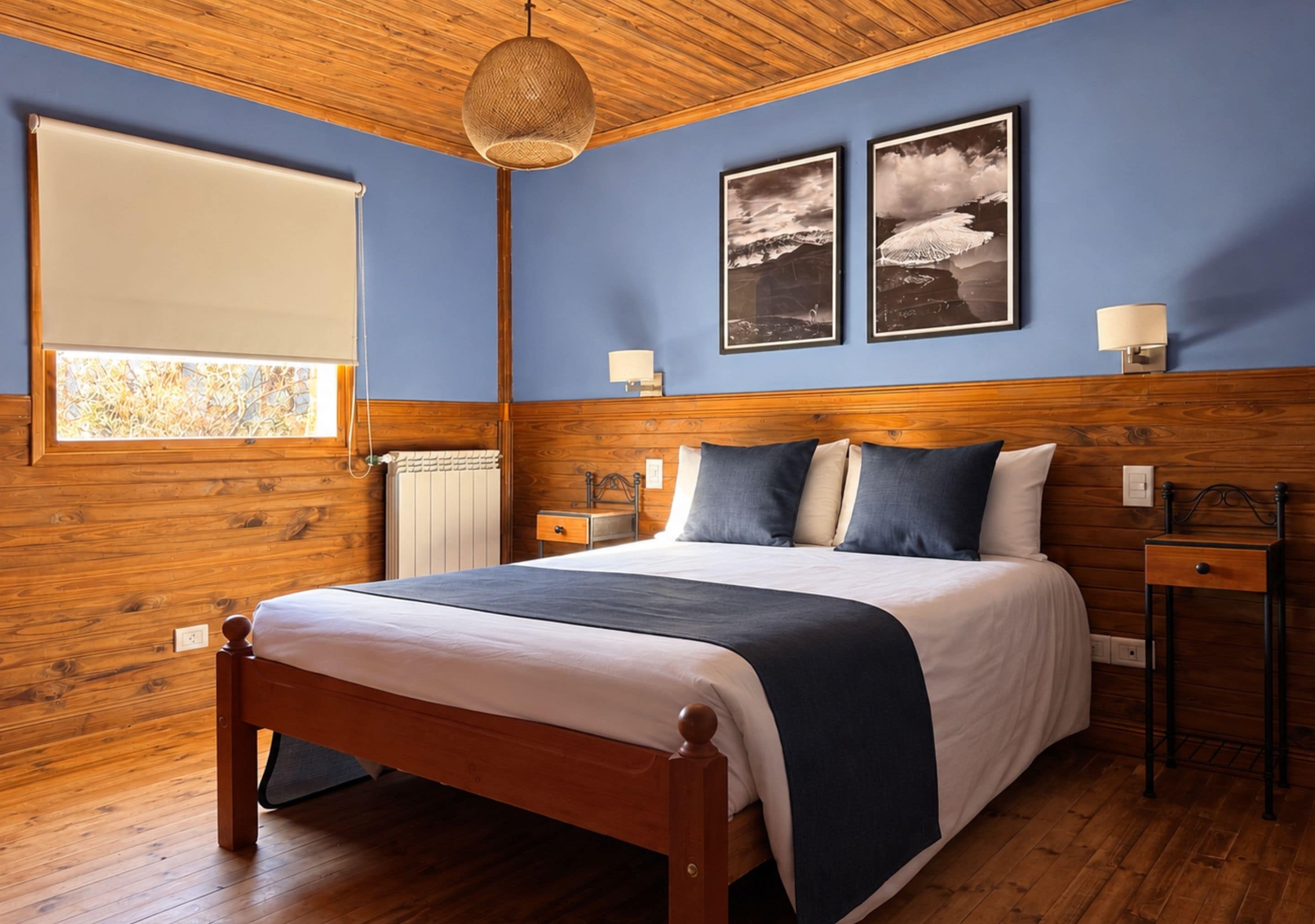 A bedroom with a wooden headboard, a bed with a white and dark blue bedspread, two bedside lamps, and two framed pictures on the wall.