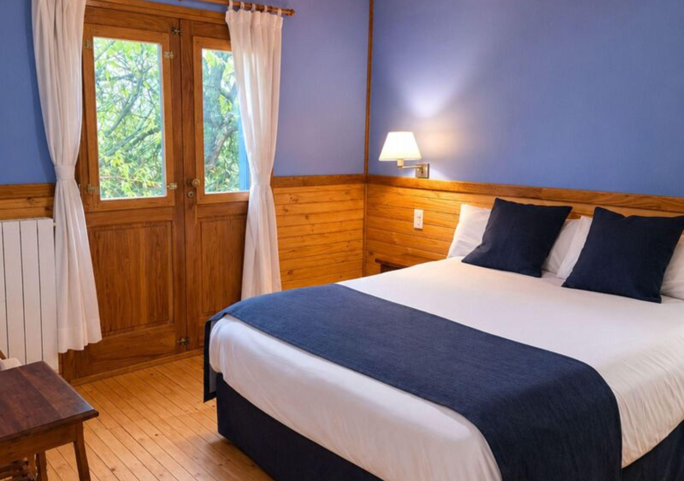 A bedroom with a wooden headboard, a bed with white and dark blue bedding, a bedside table, and a window with curtains.