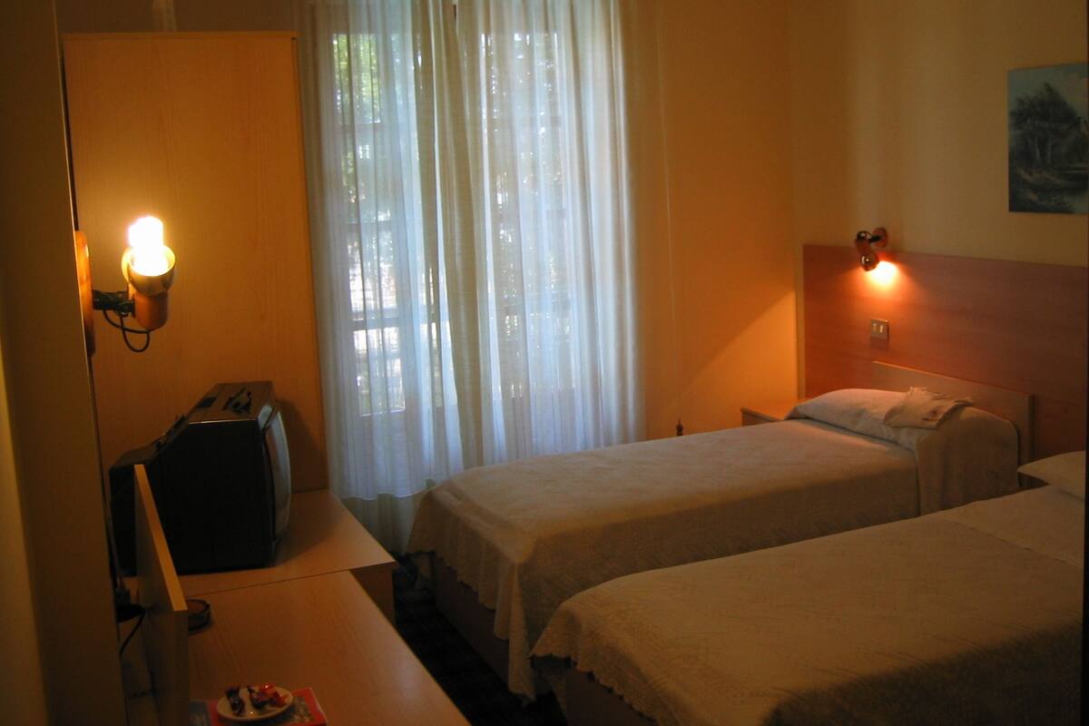 A hotel room with two beds, a desk, a chair, a suitcase, and a window with curtains.