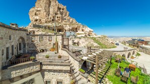 A stone building with a large rock formation in the background, outdoor seating area, and a clear blue sky. - Kale Konak (Nevsehir)