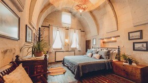 A cozy bedroom with a large bed, a wooden dresser, and a desk with a chair. There are two windows with curtains, a chandelier, and framed pictures on the walls. - Kale Konak (Nevsehir)