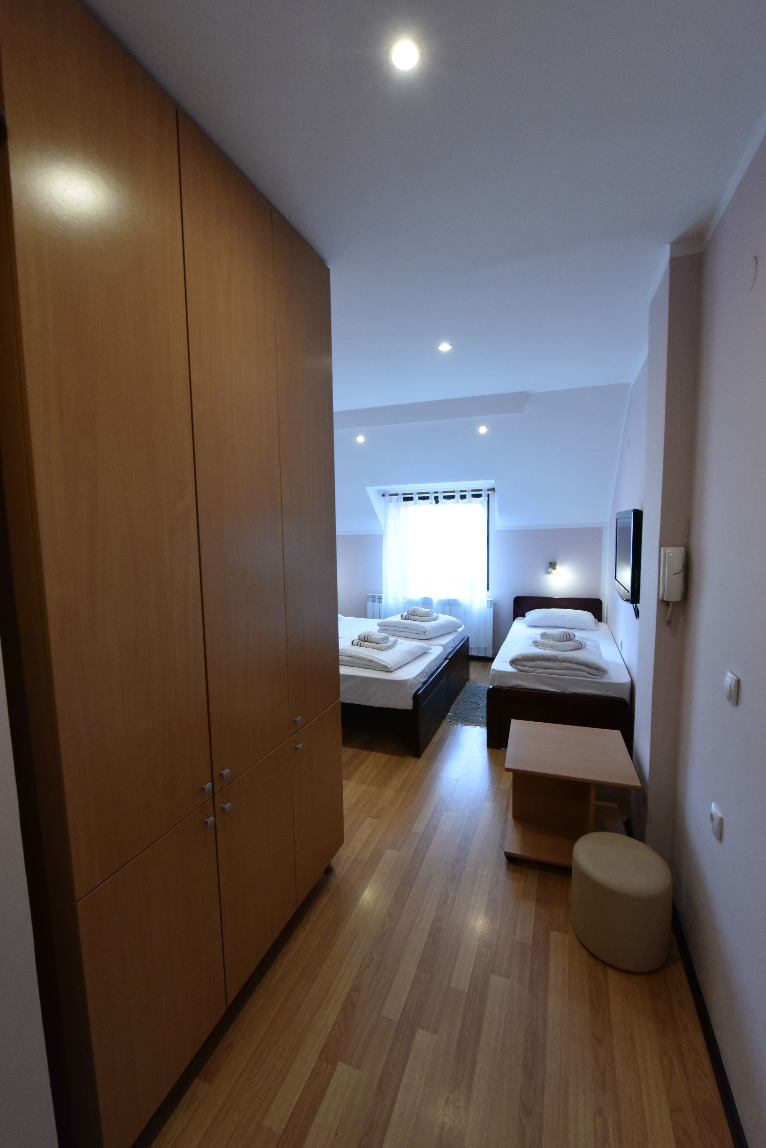 Une chambre d’hôtel moderne avec une armoire en bois, un lit doté d’une tête de lit blanche, une petite table avec un coussin et un téléviseur fixé au mur.