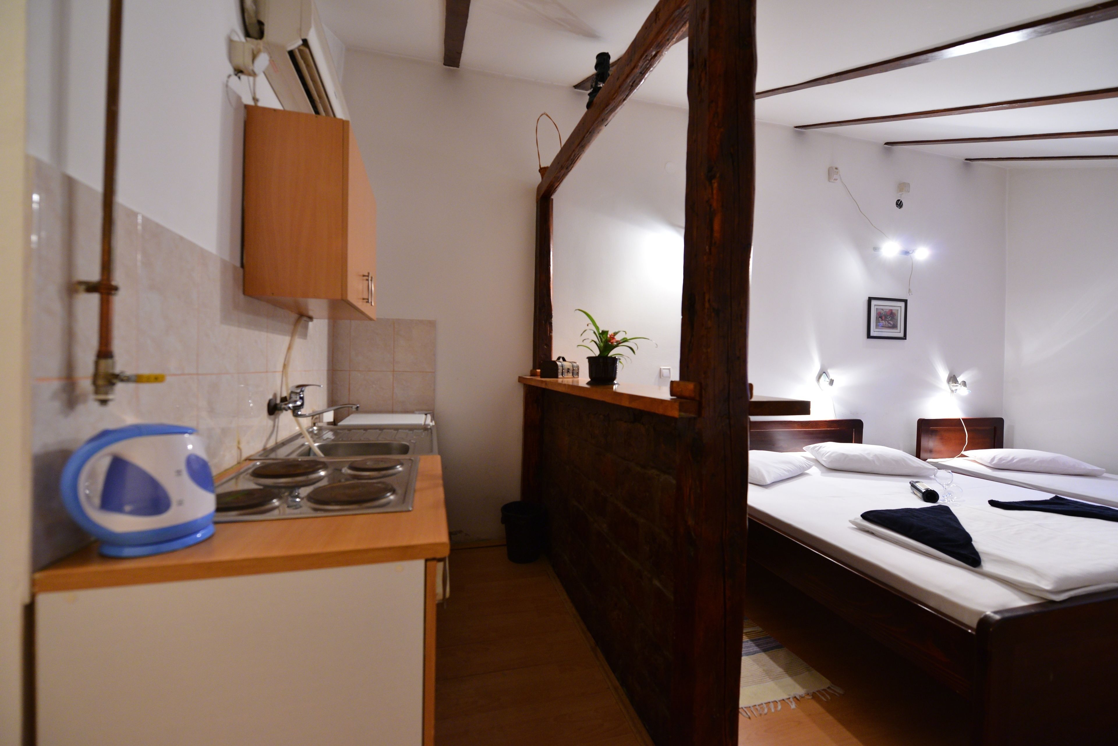 A compact hotel room with a kitchenette, a bed with two pillows, a bedside table, and a small potted plant.
