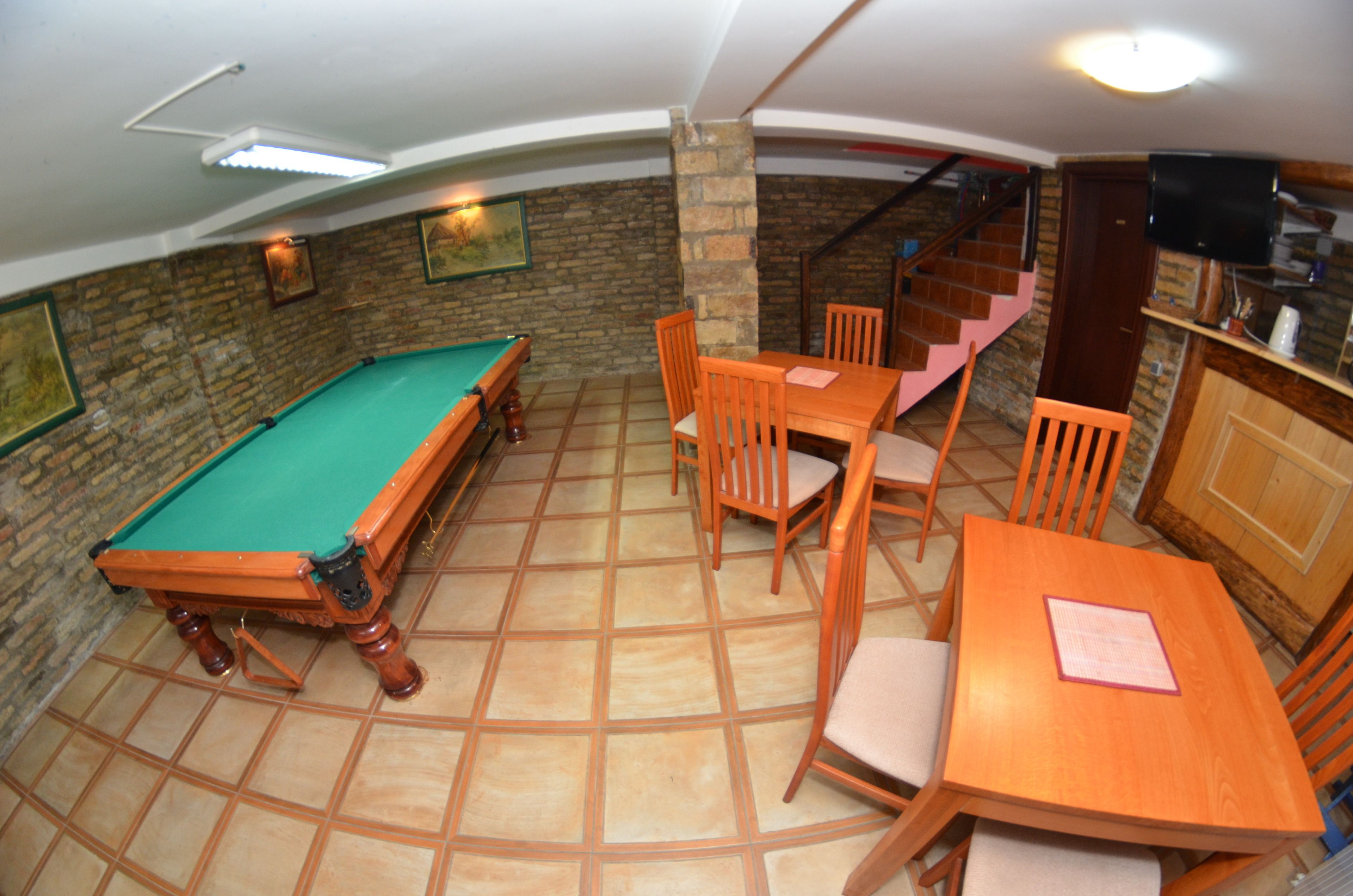 Una mesa de billar con sillas, una mesa de madera y una escalera en una habitación con suelo de baldosas y paredes de ladrillo.