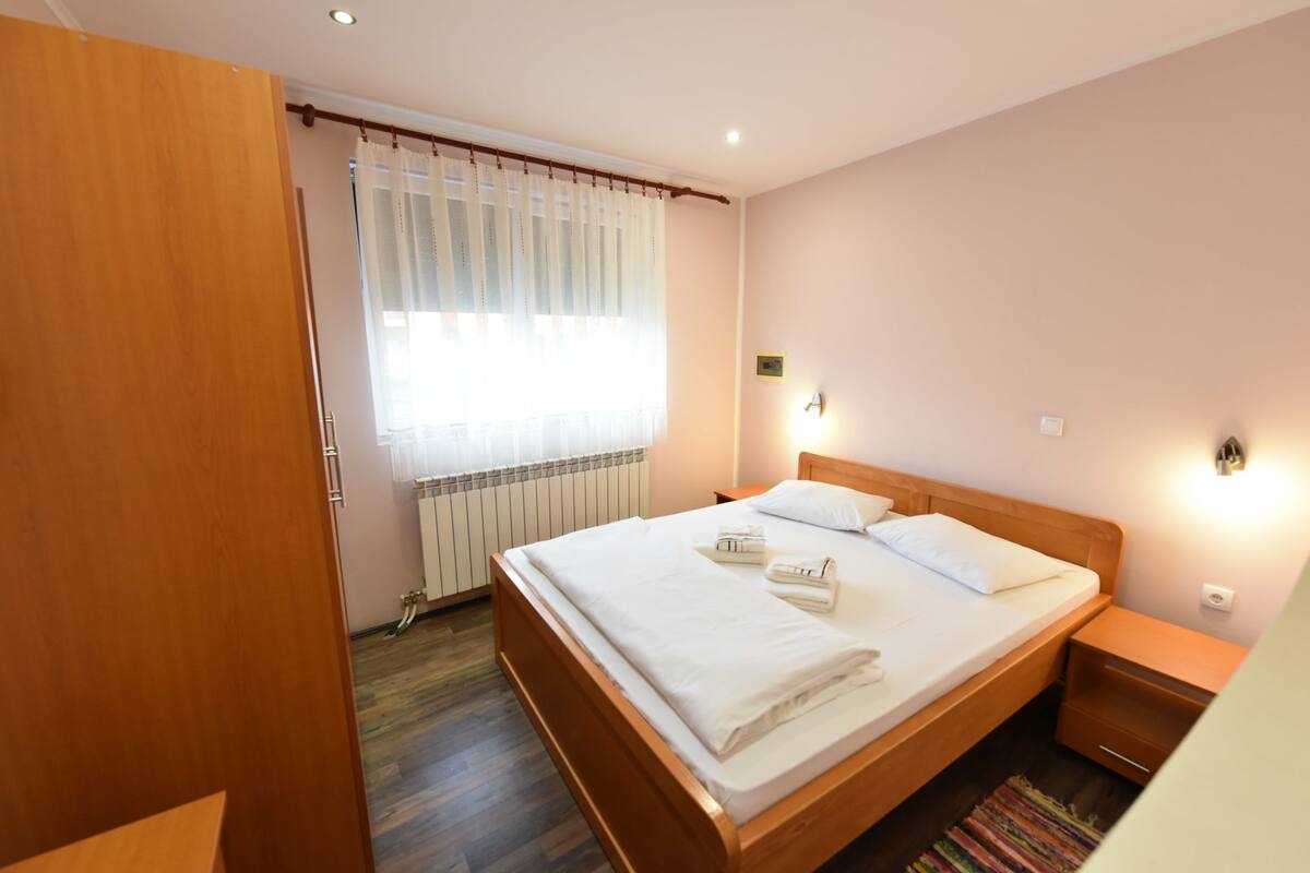 A neatly made bed with white linens and towels, a wooden headboard, a bedside table with a lamp, and a window with sheer curtains.