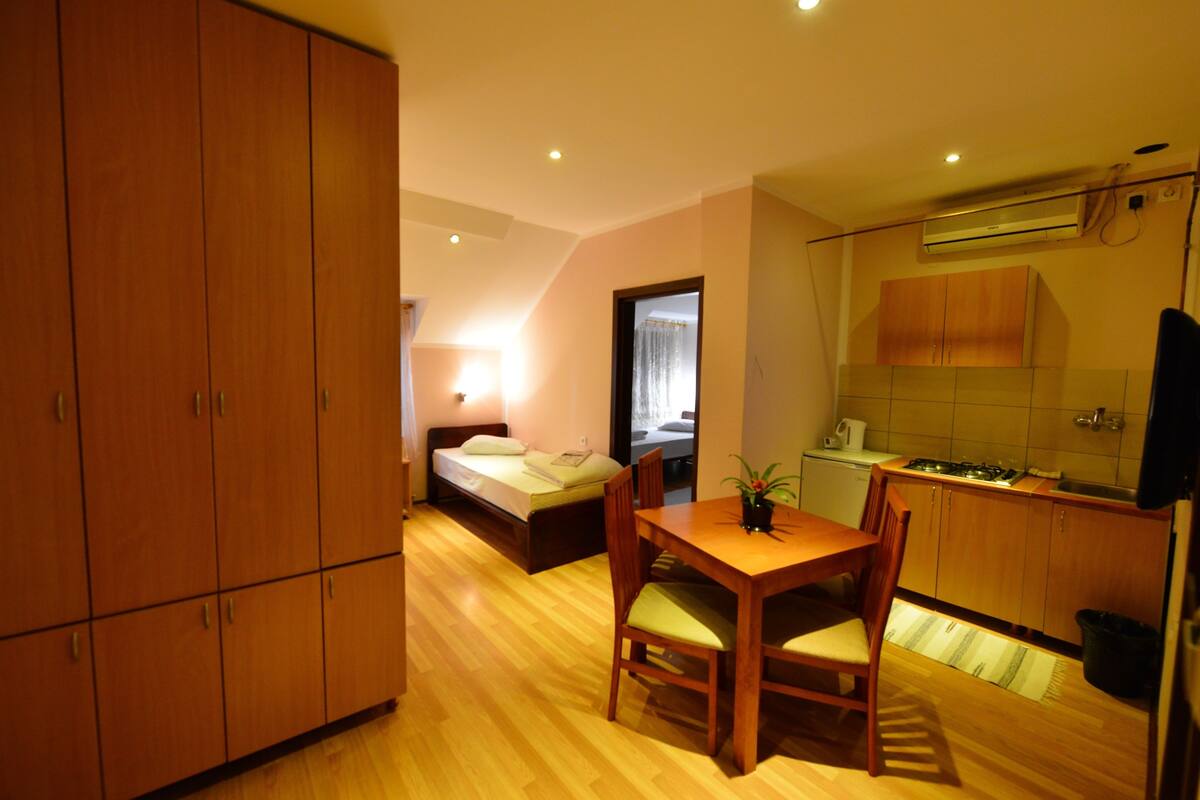 A compact room with a bed, a small dining table with chairs, a kitchenette, and wooden cabinets.