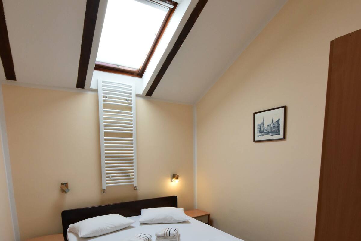 A bedroom with a skylight, a bed with white linens, a wooden headboard, a bedside table, and a small bedside lamp.