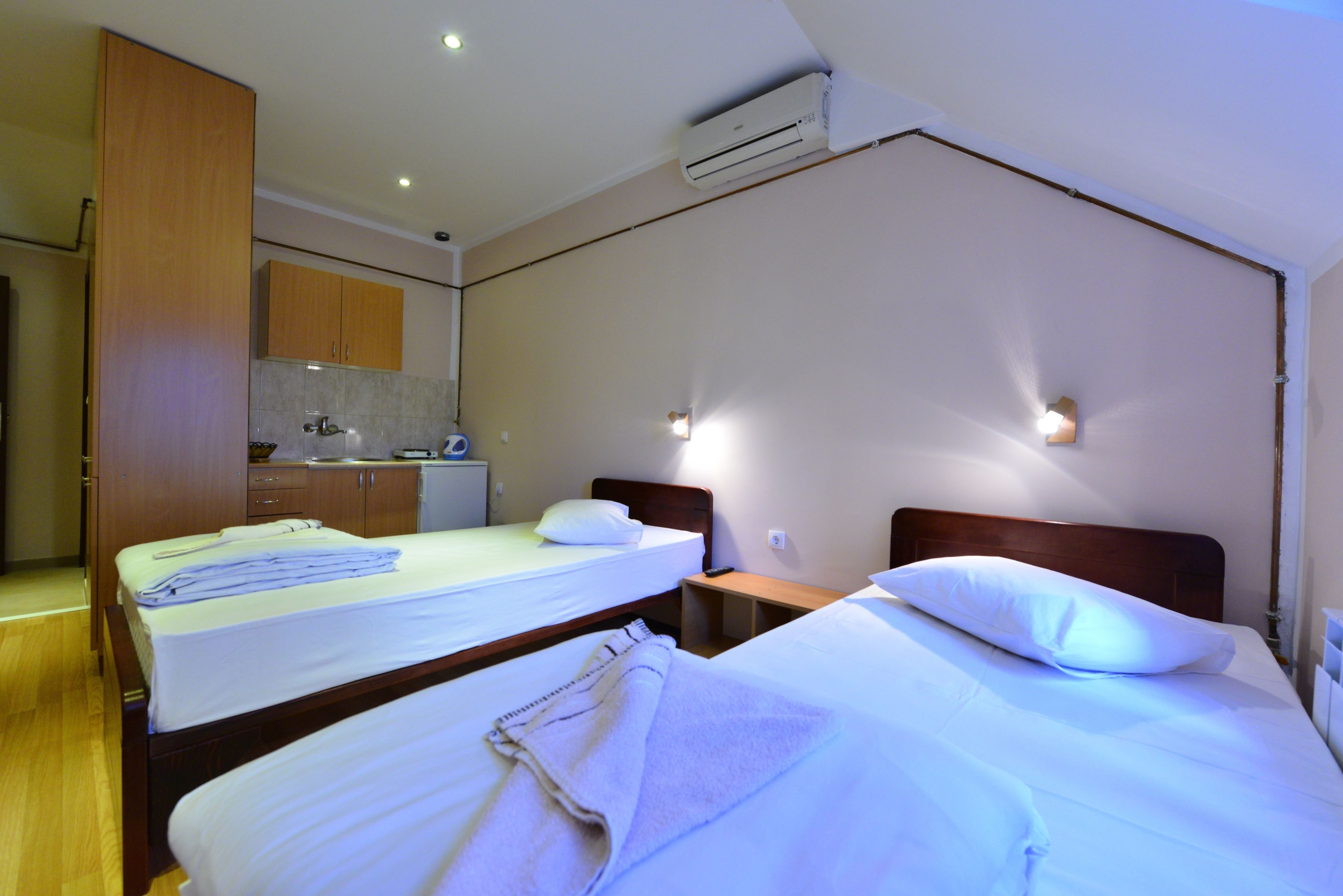 Une chambre d’hôtel avec deux lits, une armoire en bois, un petit coin cuisine et un climatiseur fixé au mur.