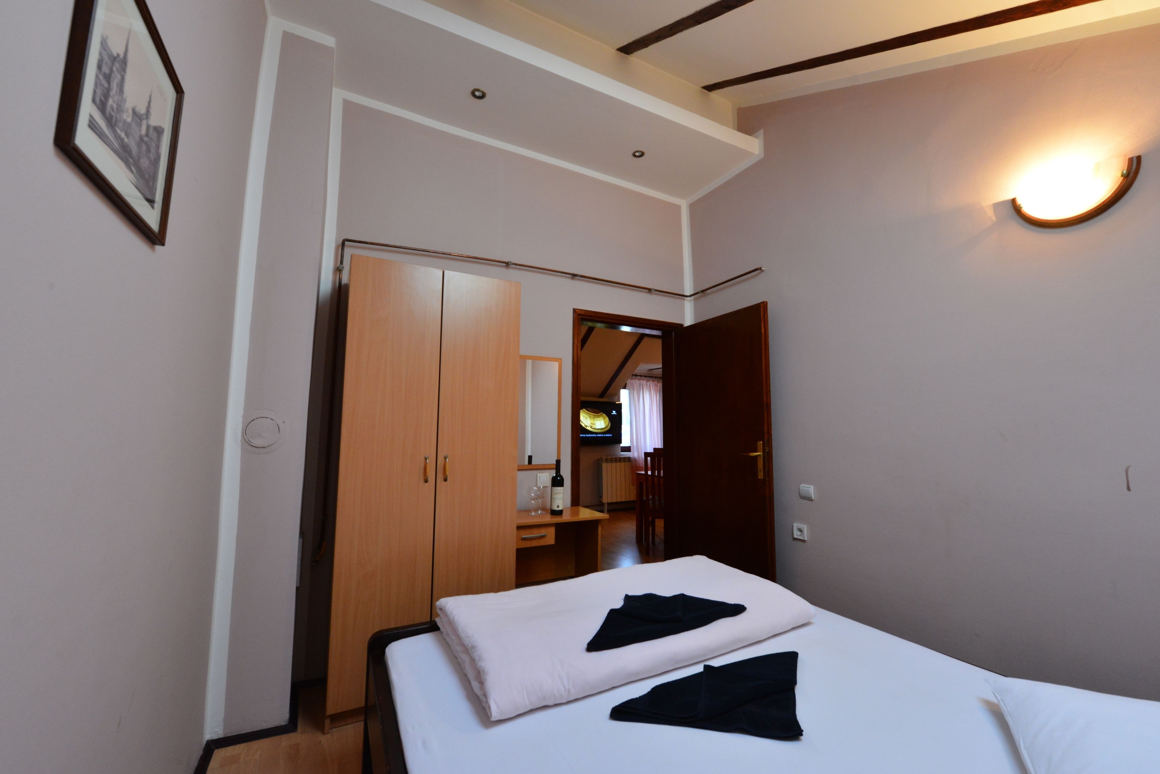 Quarto de hotel com cama, guarda-roupa, escrivaninha com cadeira, televisão e luminária na parede.