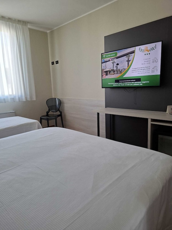 A hotel room with two beds, a TV mounted on the wall displaying hotel information, a chair, and a window with curtains. - Orzihotel (Orzivecchi)