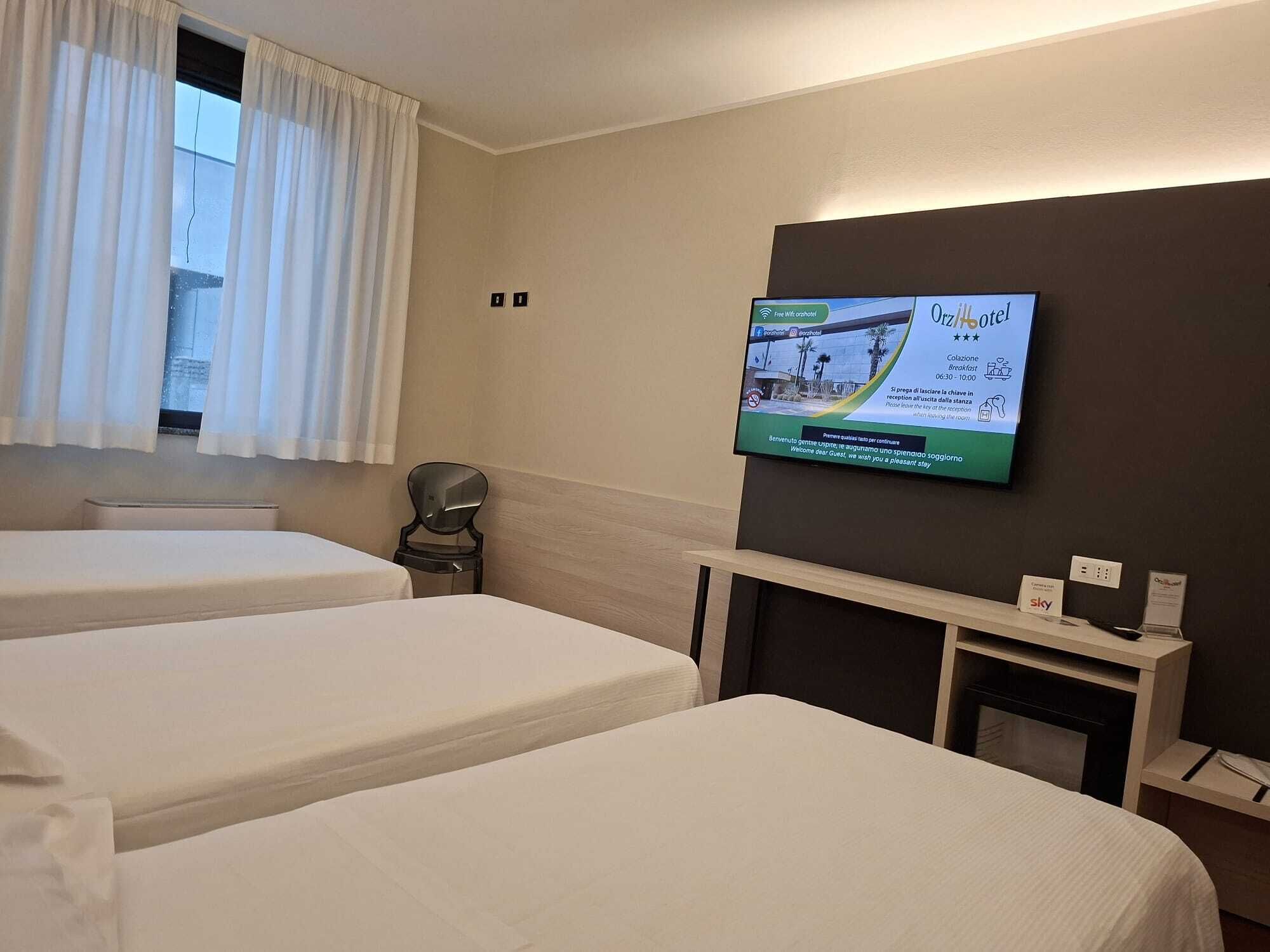 Habitación de hotel con dos camas, un televisor fijado a la pared mostrando un mensaje de bienvenida y una mesita con una silla.