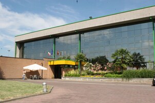Un edificio moderno con una amplia fachada de vidrio, una entrada de ladrillo y un área ajardinada con plantas y un espacio de descanso.