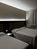 Habitación de hotel con dos camas individuales, cabecero con panel de luz, mesita de noche con teléfono y cortina en la ventana.