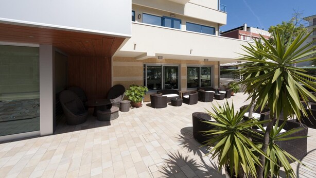 An outdoor patio area with wicker furniture, potted plants, and a building with multiple floors and balconies. - Hotel Miami (Jesolo)