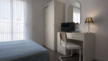 A hotel room with a bed, a desk with a computer, a chair, a lamp, a wardrobe, and a mirror.