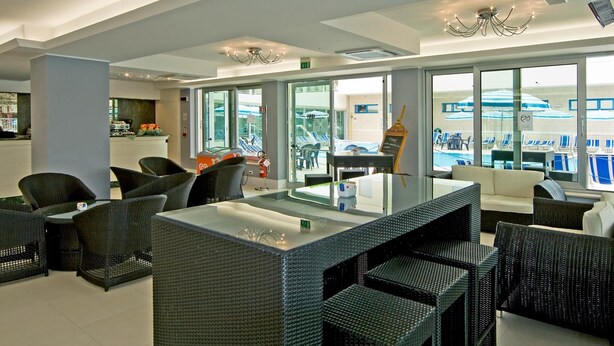 A modern hotel lobby with wicker furniture, a glass-top bar, and large windows offering views of the pool and outdoor seating. - Hotel Miami (Jesolo)