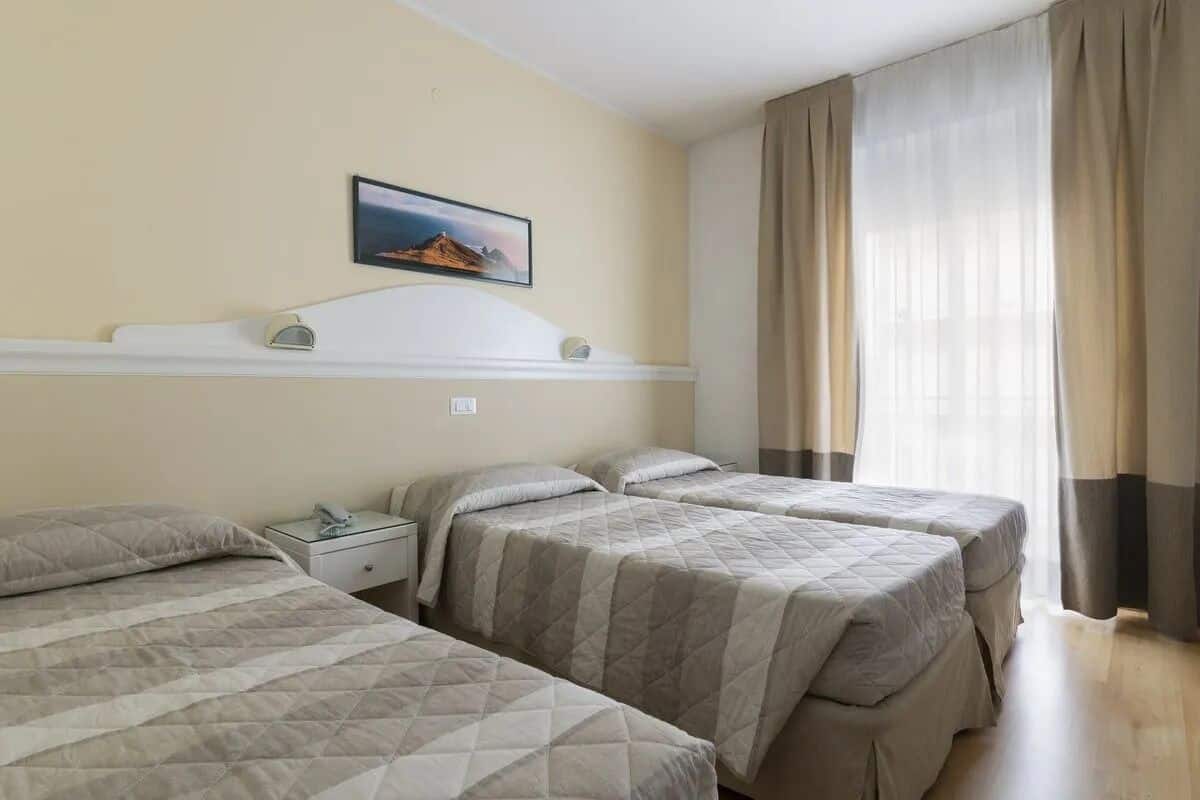 A hotel room with three beds, a bedside table with a lamp, a mounted picture on the wall, and a window with curtains.