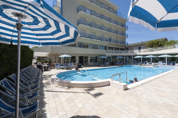 A pool area with blue and white striped umbrellas, lounge chairs, and a building with balconies. - Hotel Miami (Jesolo)