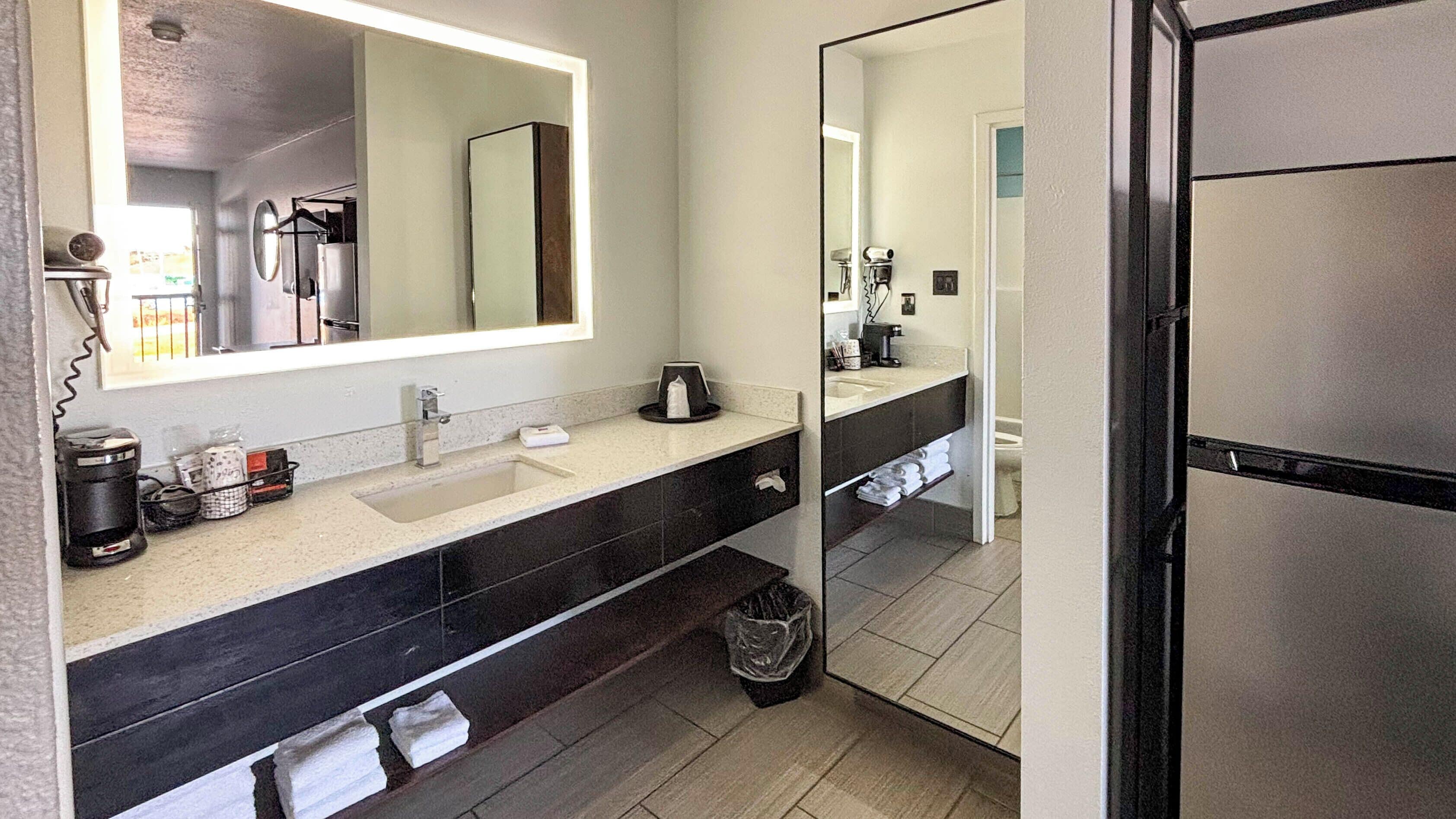 A modern bathroom with a large mirror, a sink, and a toilet. There is a trash bin and a towel rack.