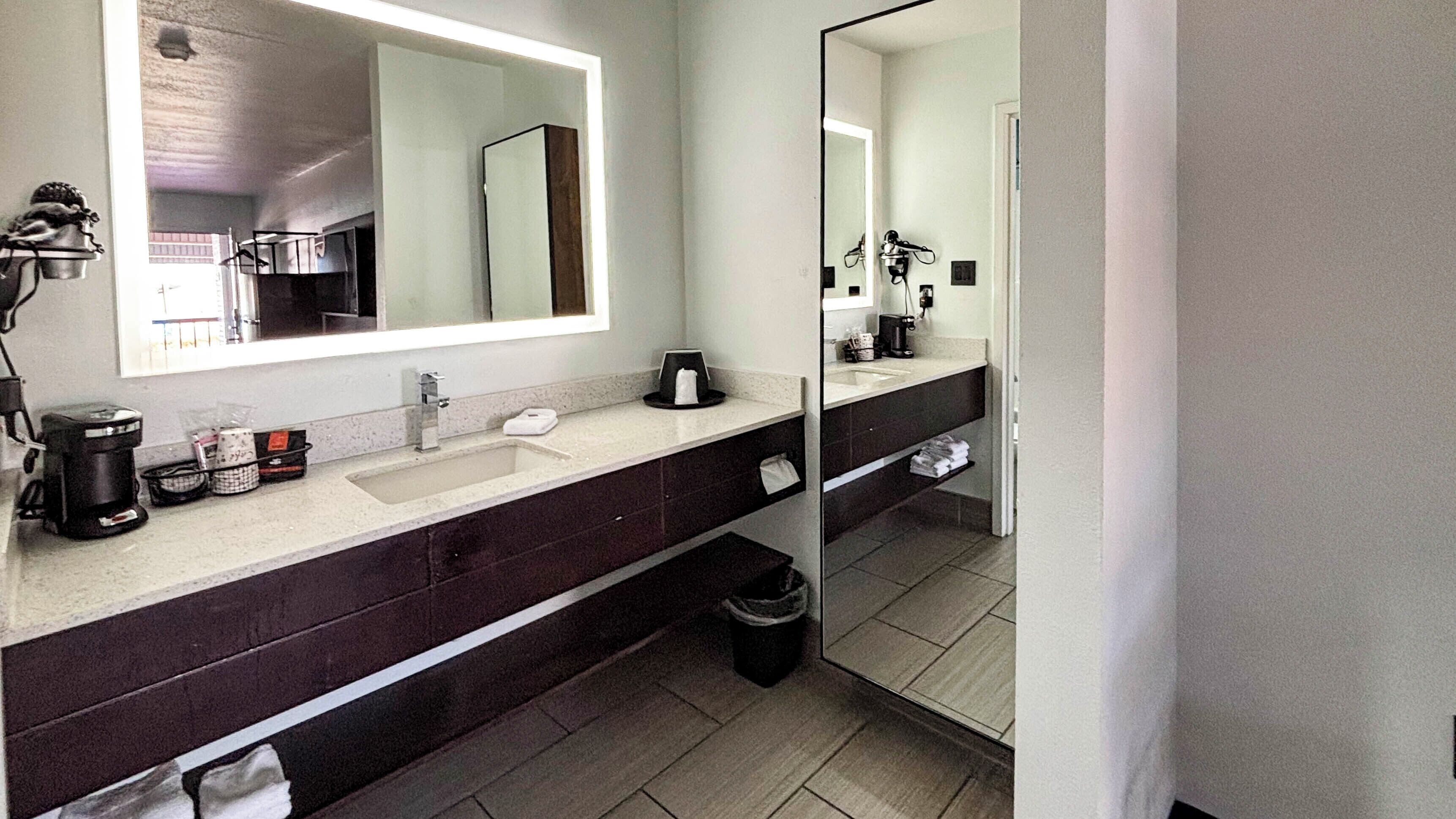 A modern bathroom with a large mirror, a sink, a coffee maker, and a trash bin.