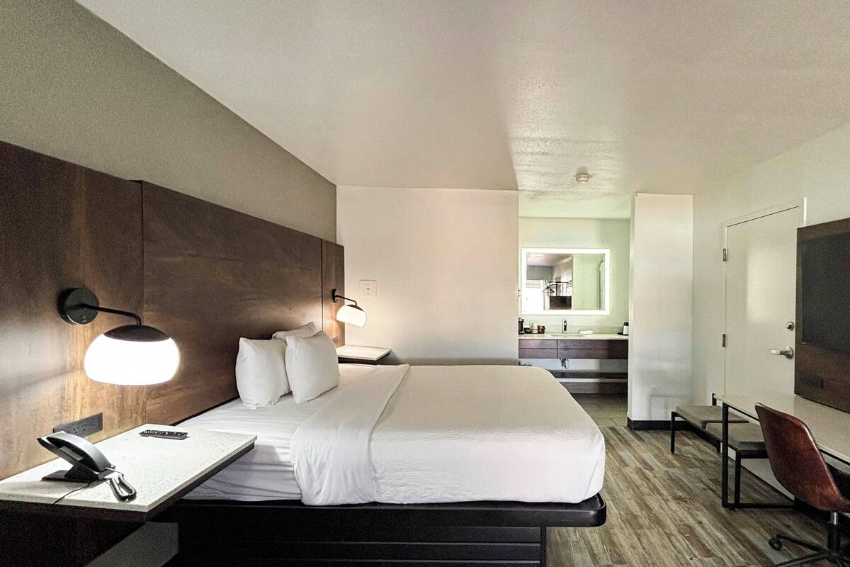 Ett hotellrum med en säng, ett sängbord, en lampa, ett skrivbord, en stol och utsikt över badrummet och köket.