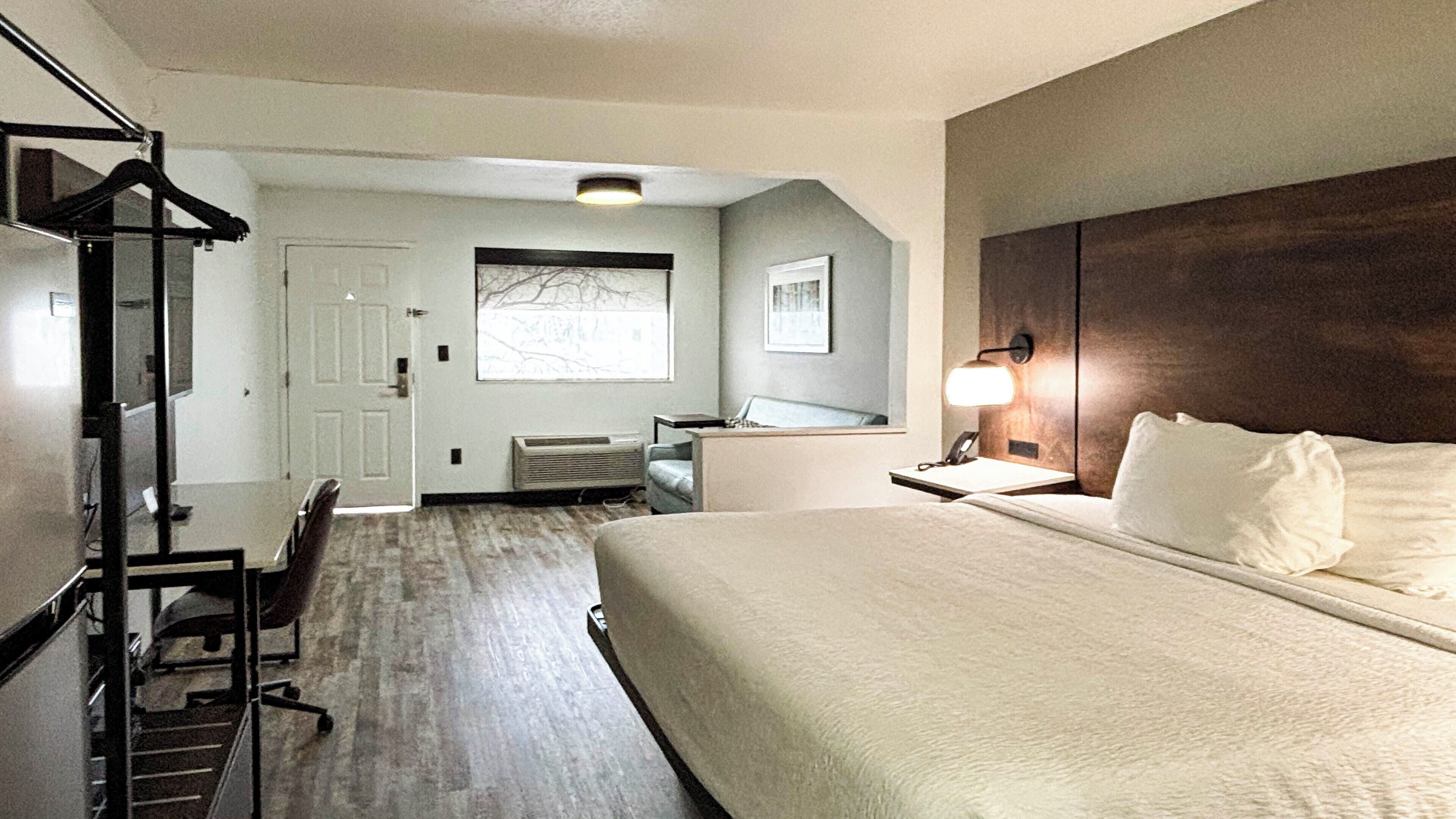 Une chambre d’hôtel avec un grand lit, un bureau avec une chaise, un porte-manteau, une fenêtre donnant sur un paysage agréable et une lampe fixée au mur.