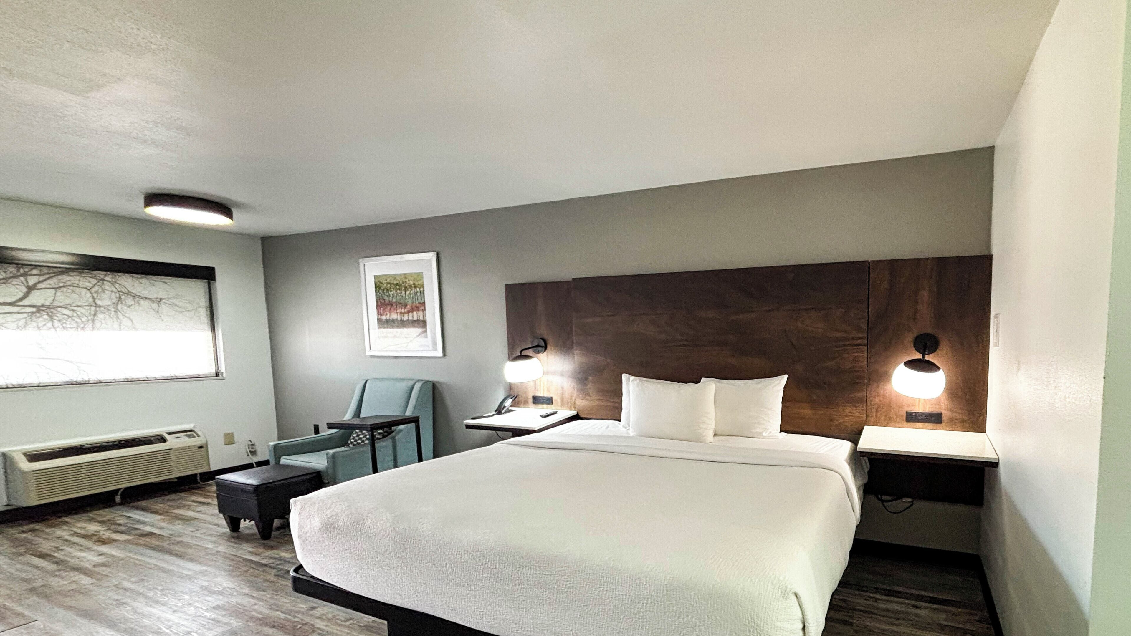 A hotel room with a large bed, a desk, a chair, a nightstand, a wall-mounted lamp, and a framed picture on the wall.