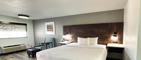 A hotel room with a large bed, a desk, a chair, a nightstand, a wall-mounted lamp, and a framed picture on the wall.