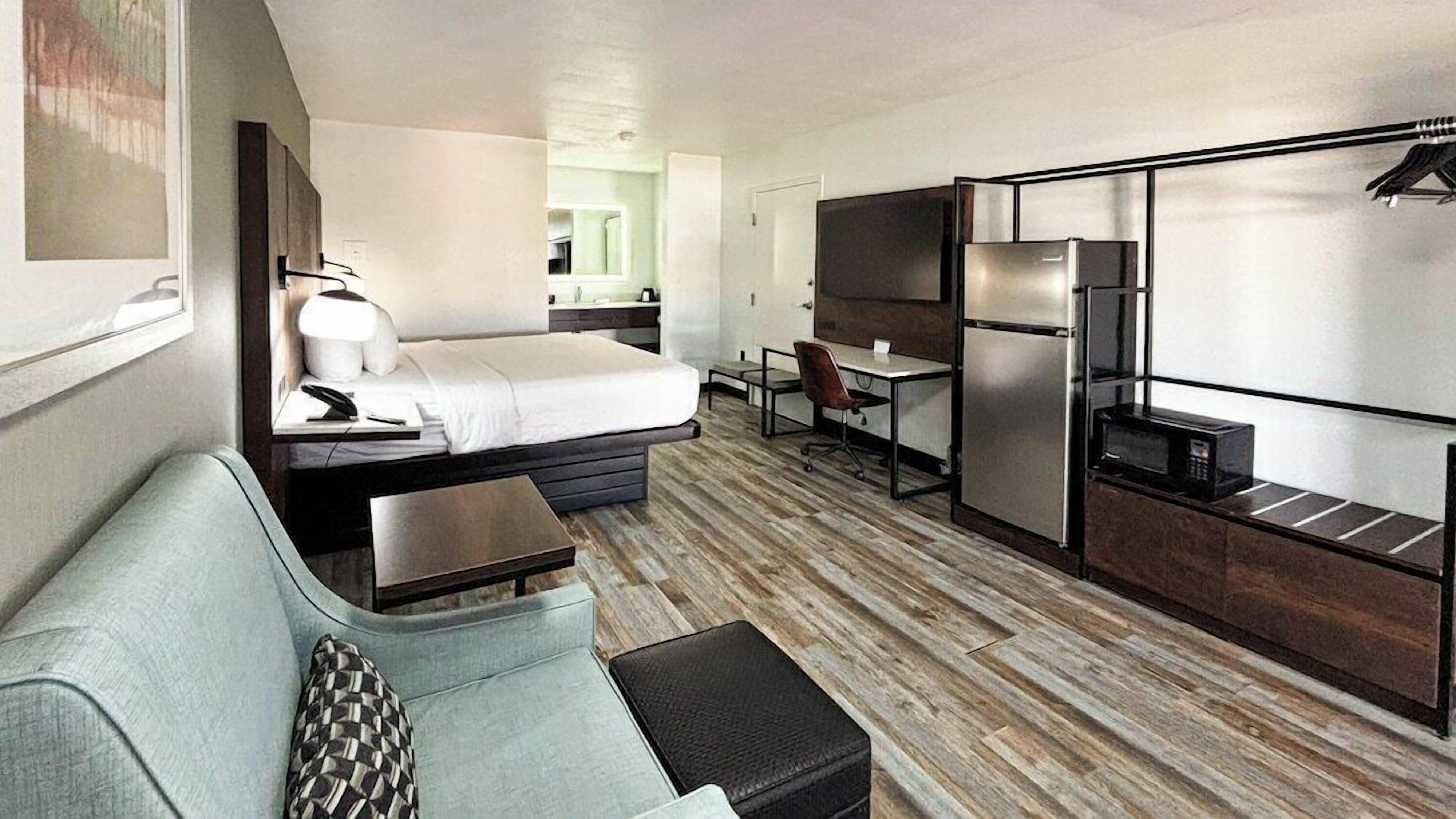 A compact hotel room with a bed, a small sofa, a desk, a chair, and a kitchenette.