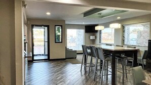 Cocina moderna con piso de madera, taburetes de bar y una mesa de comedor. Hay dos ventanas con vistas al exterior.