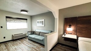Una habitación pequeña y acogedora con un sofá azul claro, un cuadro en la pared, una ventana con vistas a árboles desnudos, un calefactor en la pared y una lámpara de noche.