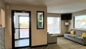 A hotel lobby with a reception desk, a sofa with cushions, a television mounted on the wall, and a large window with a view of the outside. - Studio 6 Suites – Richland, MS / Jackson (Richland)