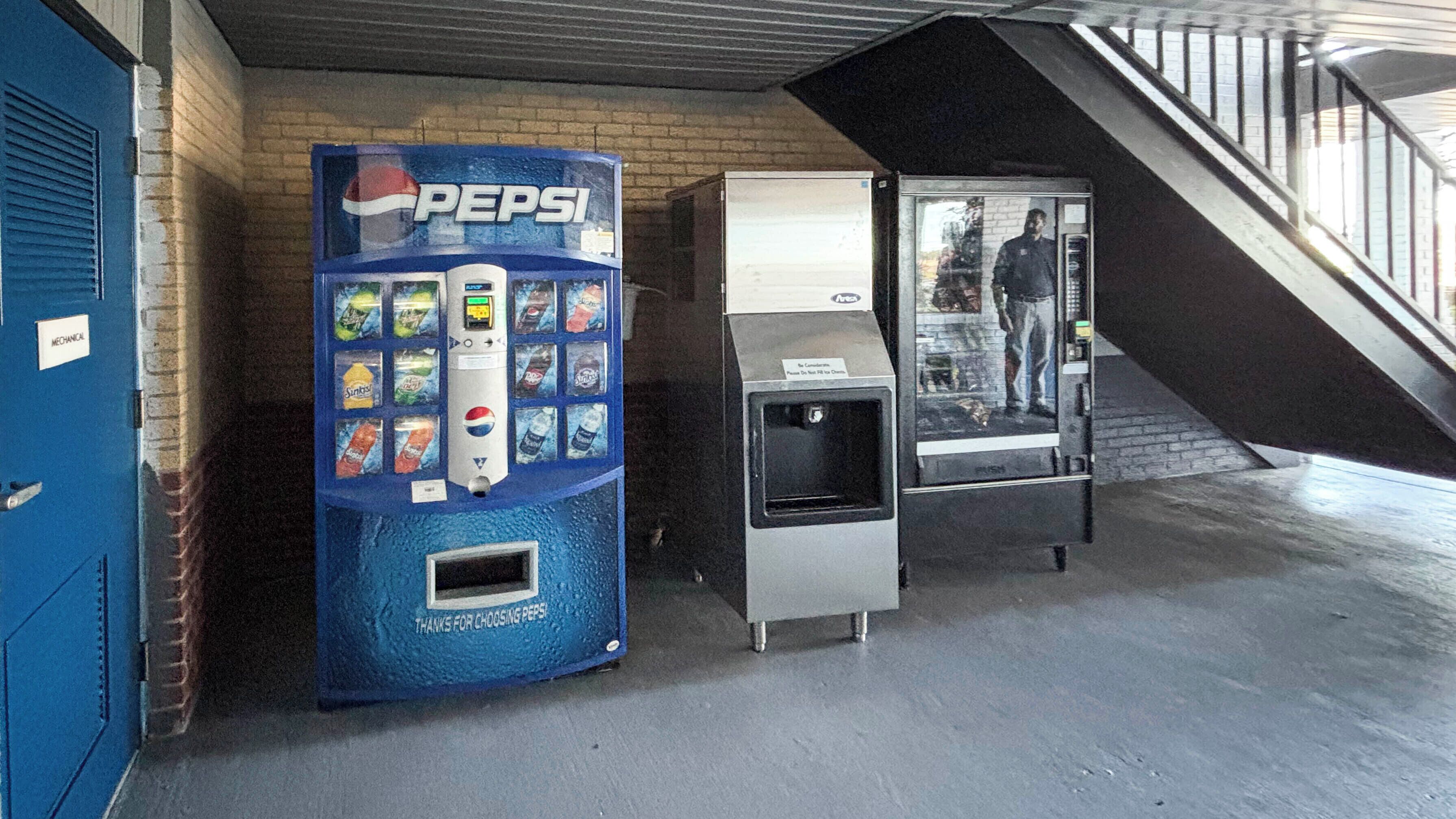 Een automaat met Pepsi en een waterdispenser in een industriële omgeving, met een bakstenen muur en een metalen trap.