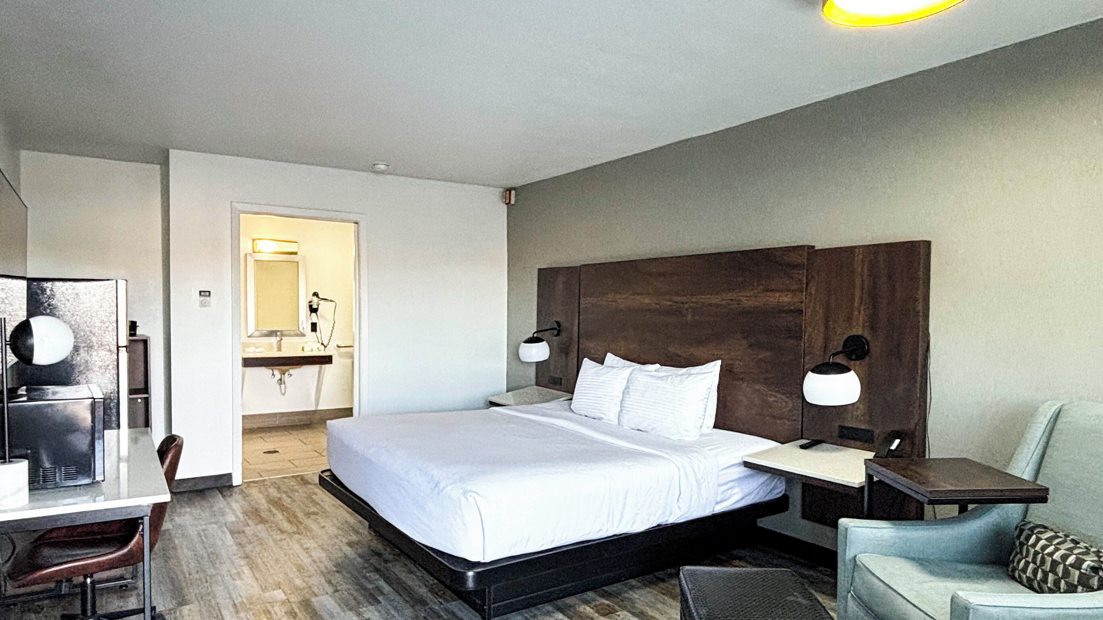 A hotel room with a bed, bedside tables, a desk, a chair, and a bathroom visible through an open door.