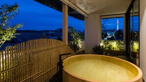 Een traditioneel Japans onsen met een houten bad, een bamboebegraafing en uitzicht op een stadsgezicht met een toren.