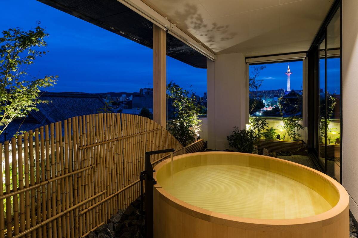 Un onsen japonés tradicional con una bañera de madera, valla de bambú y vista a un paisaje urbano que incluye una torre.